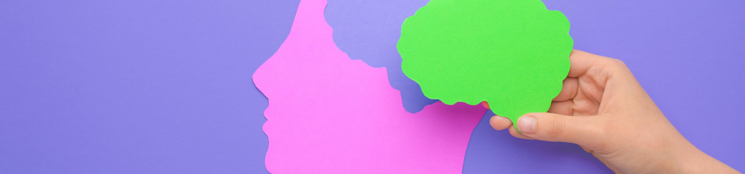 A hand holding a paper cutout of a human brain over the paper cutout of a human head with a pastel purple color background